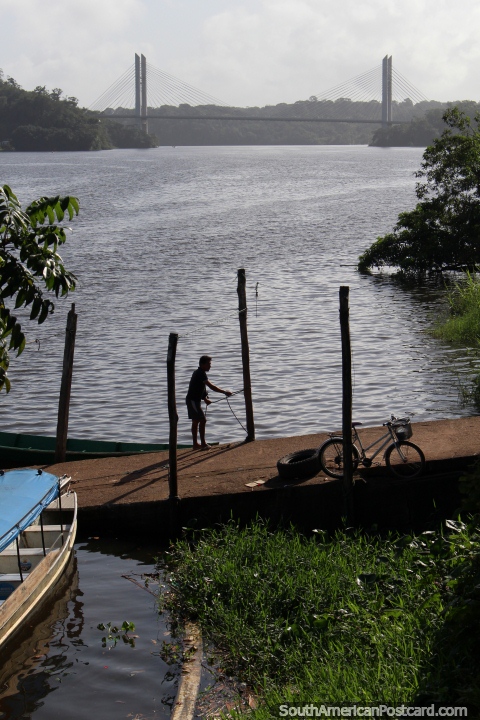O homem amarra o seu barco junto de uma rampa no rio em Oiapoque, a ponte a Guiana Francesa na distância. (480x720px). Brasil, América do Sul.