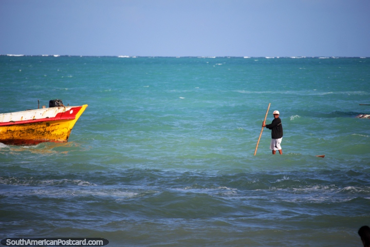 Man standing on a board in the water navigates from his boat to the shore with a stick in Maragogi. (720x480px). Brazil, South America.