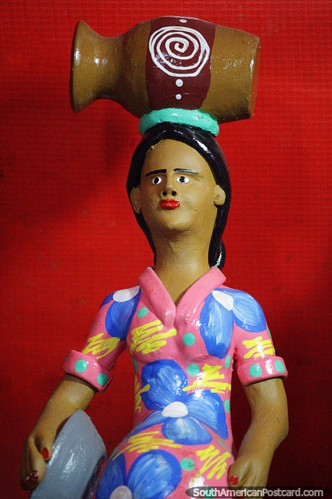 Woman in a pink and blue floral dress has an urn on her head, ceramics in Porto Velho. (480x720px). Brazil, South America.