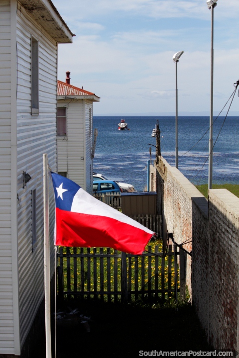 A bandeira chilena voa com o mar a vista em volta de casas na terra a margem de água em Punta Arenas. (480x720px). Chile, América do Sul.