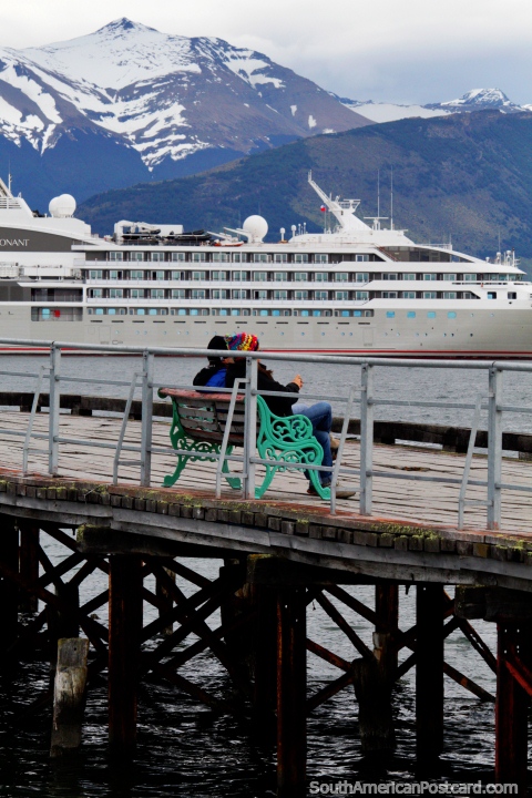 Personas sentadas en el nuevo muelle en Puerto Natales con barco distante y montañas. (480x720px). Chile, Sudamerica.