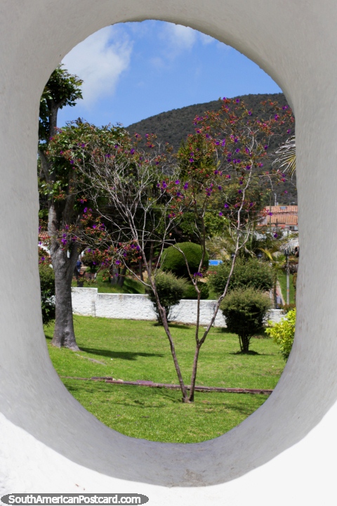 Visão por uma janela oval a árvores com flores purpúreas e gramados verdes em Guatavita. (480x720px). Colômbia, América do Sul.