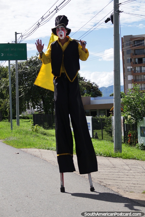 Man on stilts wearing makeup, standing on the roadside waving a flag in Girardot. (480x720px). Colombia, South America.