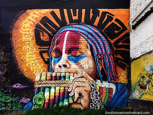 Hombre indígena con tocados y pintura facial a juego sopla tubos de viento de colores del arco iris, arte callejero en Pasto. (640x480px). Colombia, Sudamerica.