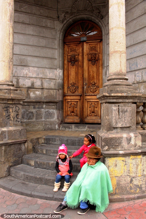 Una mujer indígena y 2 nietas sentarse fuera de un edificio de piedra en Ambato. (480x720px). Ecuador, Sudamerica.