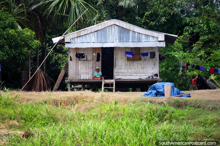 Uma mulher senta-se no pórtico da sua vida de rio de observação de casa de Amazônia de madeira vão por entre Iquitos e Santa Rosa. (720x480px). Peru, América do Sul.