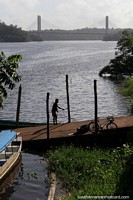 O homem amarra o seu barco junto de uma rampa no rio em Oiapoque, a ponte a Guiana Francesa na distância. Brasil, América do Sul.