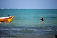 Man standing on a board in the water navigates from his boat to the shore with a stick in Maragogi. Brazil, South America.