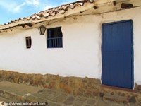 Versión más grande de Casa mona en Barichara con pared blanqueada, lámpara y puerta de madera azul.