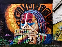 Indigenous man with matching headgear and face paint blows rainbow colored windpipes, street art in Pasto. Colombia, South America.