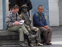A bronze man sits between 2 real men on a bench seat in Quito. Ecuador, South America.