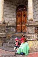 An indigenous woman and 2 granddaughters sit outside a stone building in Ambato. Ecuador, South America.