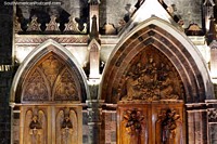 Larger version of Large arched wooden doors with intricate engraved sculptures, facade of the church in Banos at night.