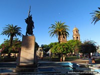 Larger version of The beautiful Plaza Constitucion in Dolores with monument, cathedral, palms and magneta leaved trees.