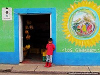 Larger version of Young boy in a red shirt and gumboots stands outside a fruit shop in Mucuchies.