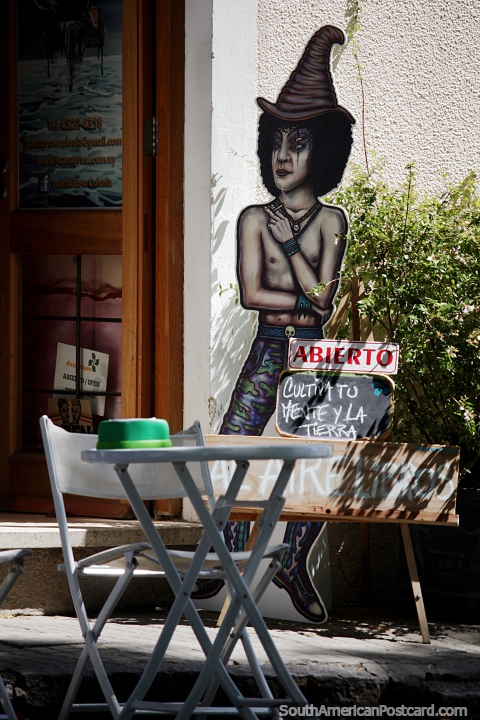 Extraa figura se encuentra fuera de una tienda con una mesa y una silla en Colonia del Sacramento. (480x720px). Uruguay, Sudamerica.